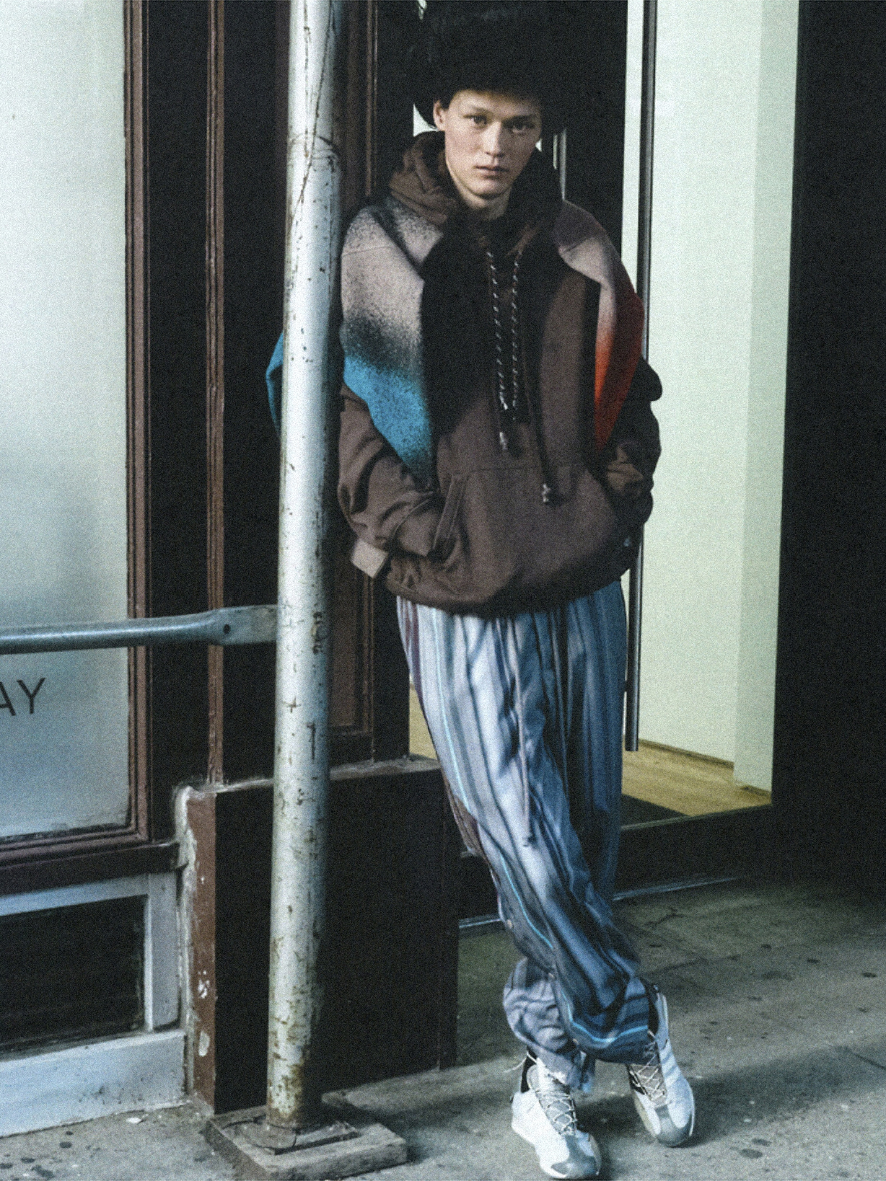 Male fashion model wearing fur hat and brown hoodie, standing against a weathered wall in an urban environment. Fashion editorial style.