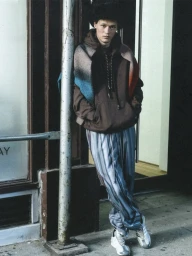 Young man leaning against a vertical metal pole wearing a brown hoodie, striped pants, and a colorful scarf draped across his shoulders.