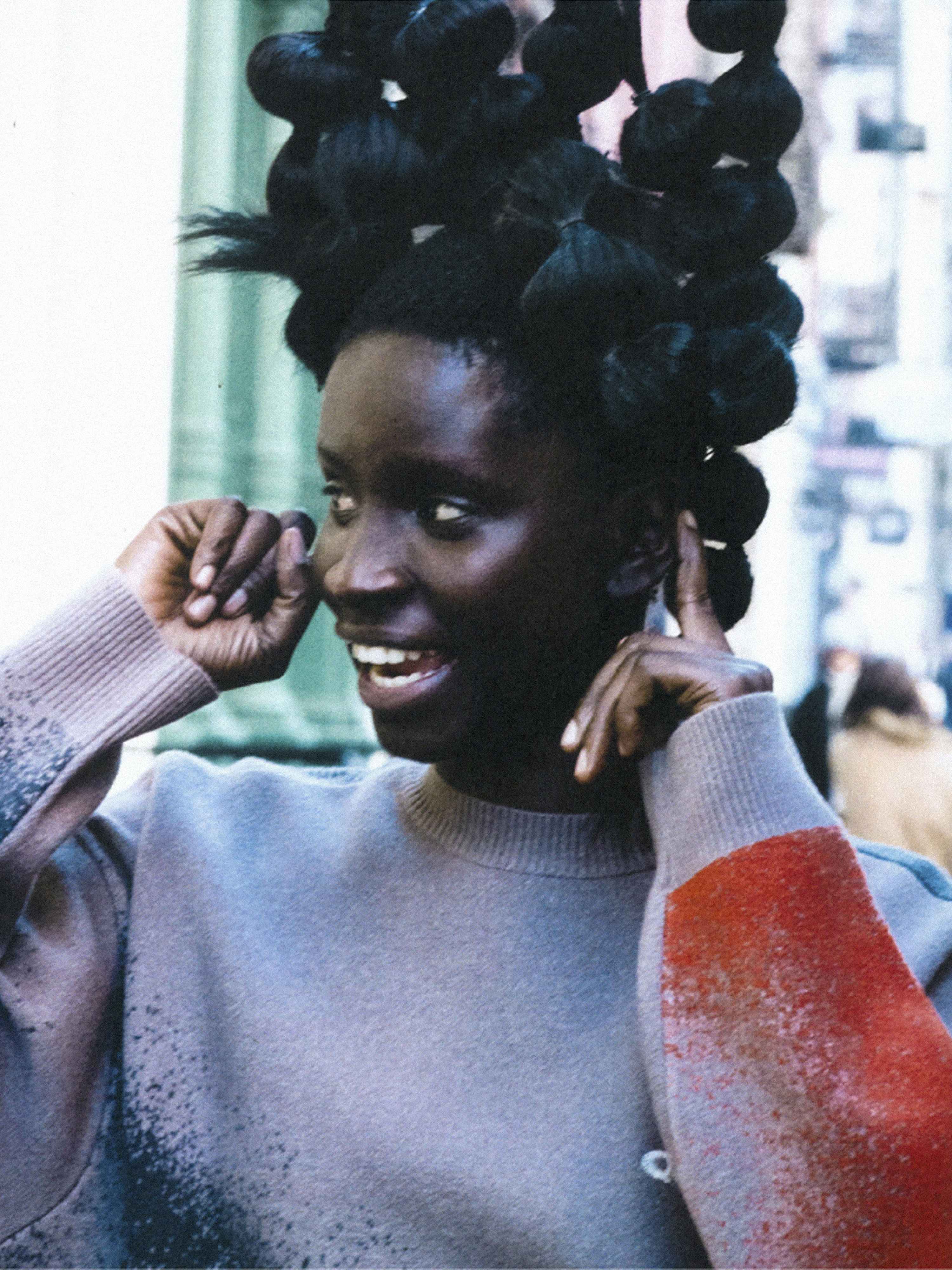 A smiling Black woman with a unique hairstyle poses outdoors. She wears a sweater with color accents against a blurred background.
