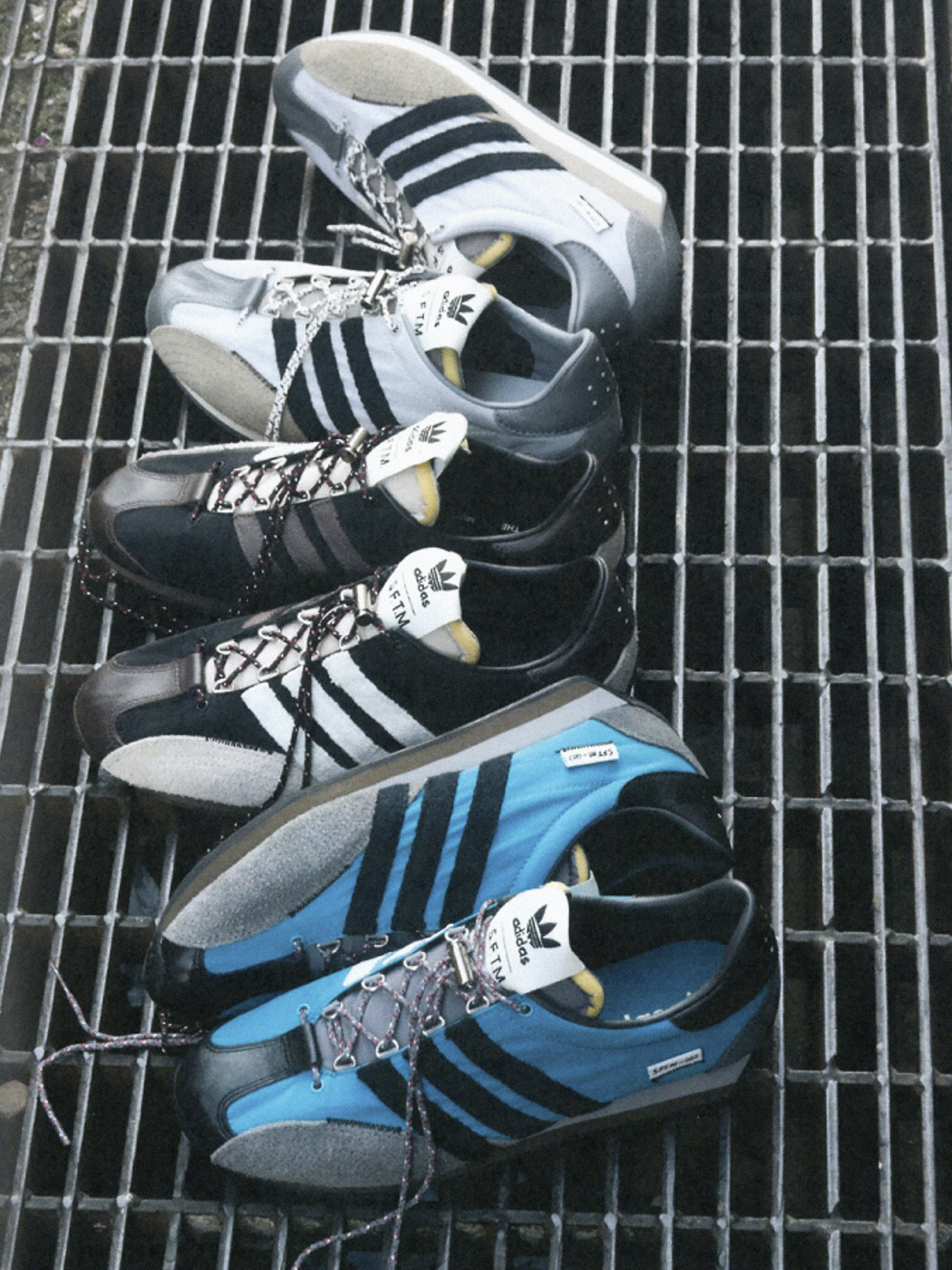 Multiple pairs of Adidas sneakers are displayed on a silver metal grate, featuring a variety of colors including white, black, and blue.