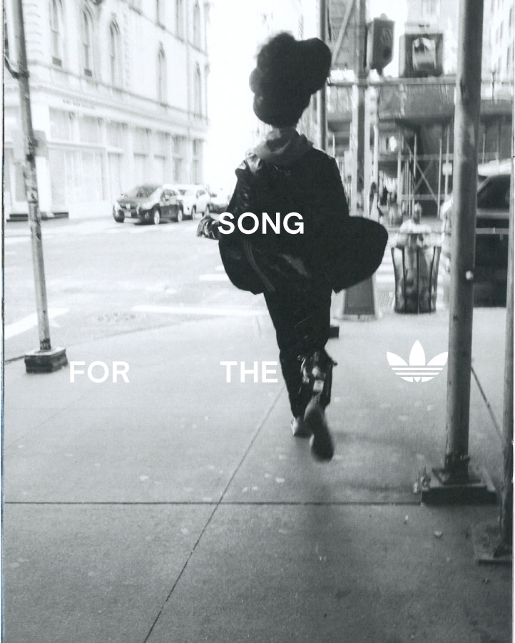 Monochrome street-style image of a person walking away on an urban sidewalk, with Adidas logo, flanked by buildings and parked cars.