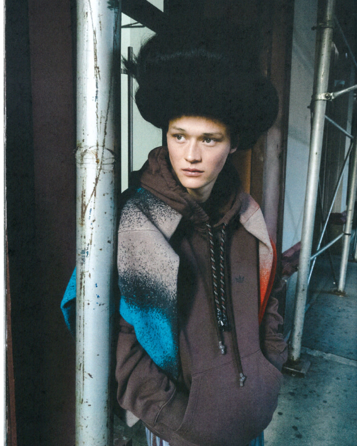 Portrait of a young adult with a unique hairstyle wearing a brown hoodie and standing outdoors against a backdrop of scaffolding and pipes.