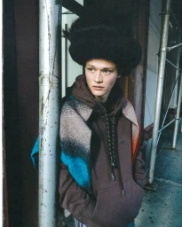 Model wearing a brown hoodie with a colourful scarf and large black hat posing near metal scaffolding poles.