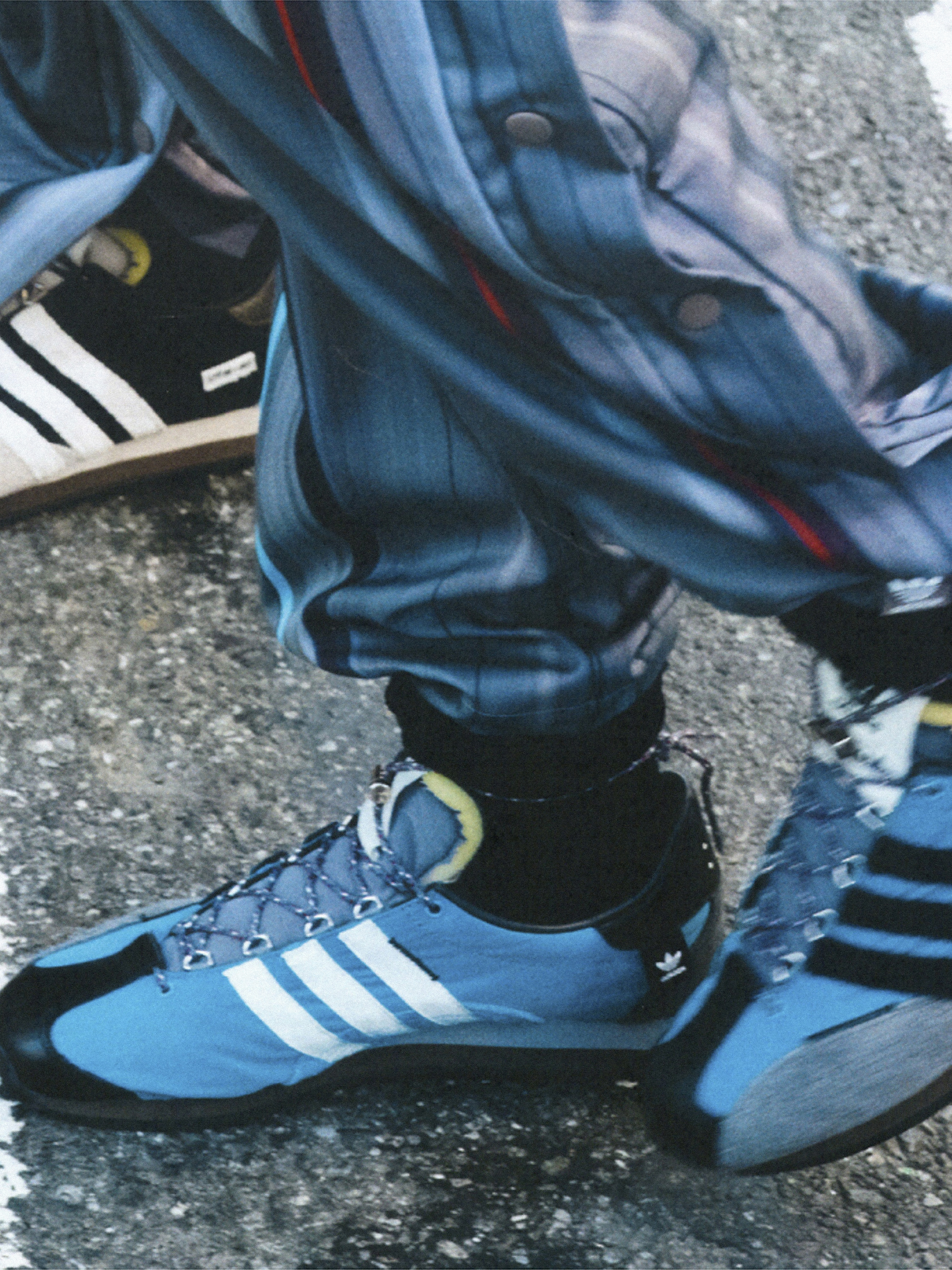 Close-up shot of a person wearing blue Adidas sneakers with white stripes, complemented by patterned pants on an asphalt surface.