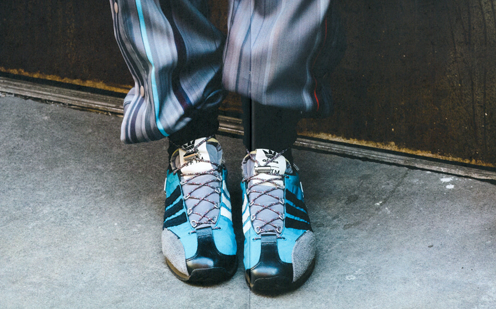 Low-angle shot of black Adidas sneakers with a blue accent, gray laces, and striped pants against a concrete backdrop.