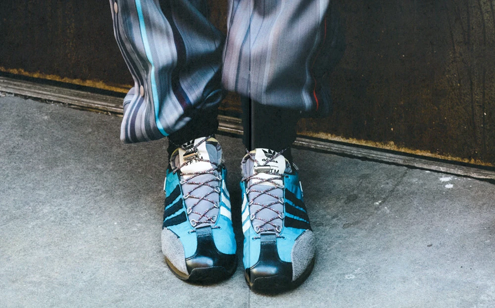 Low-angle shot of black Adidas sneakers with a blue accent, gray laces, and striped pants against a concrete backdrop.