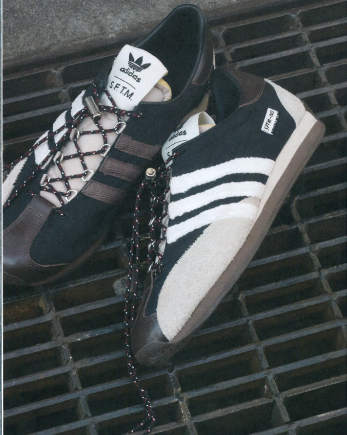 Two Adidas SFTM running shoes sit on a metal grate, one black with brown stripes and laces, the other black with white stripes.