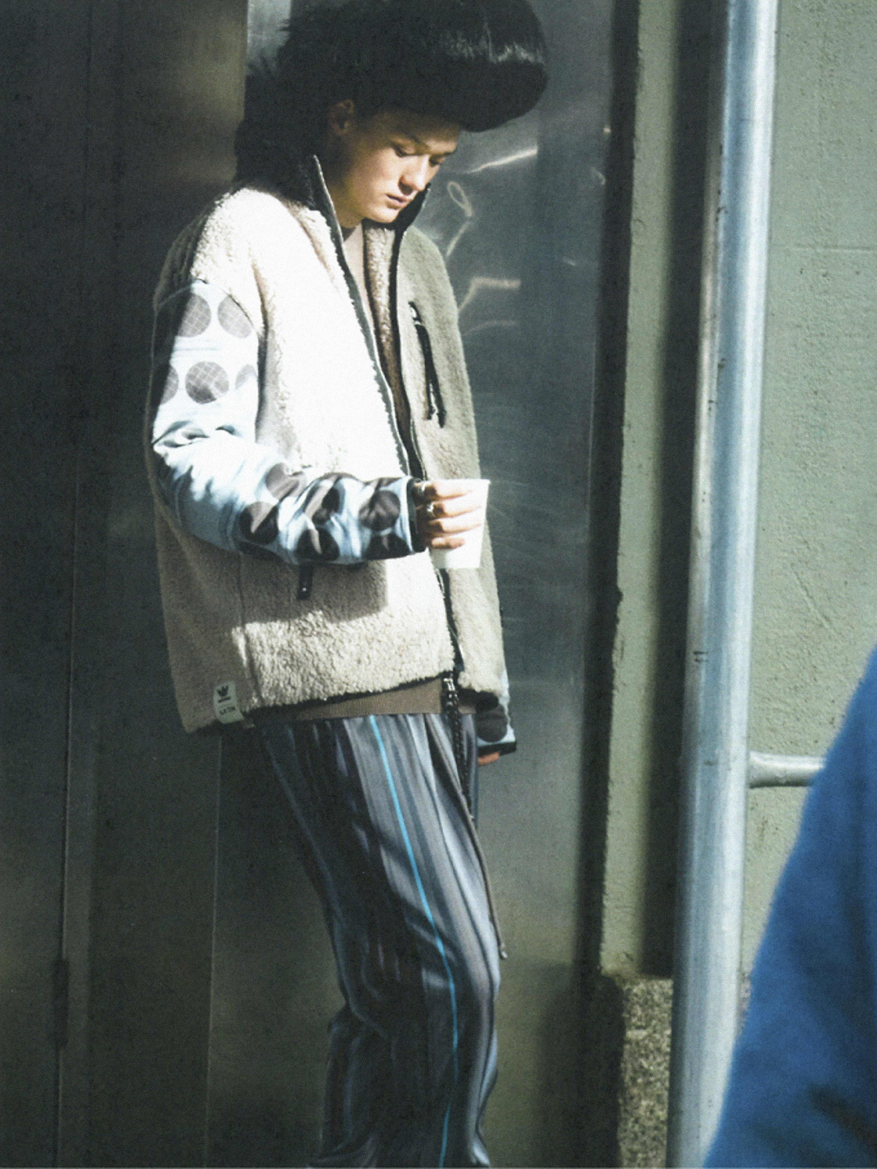 Man in oversized fur hat and patterned jacket holds cup outdoors against gray metal wall and blue railing.