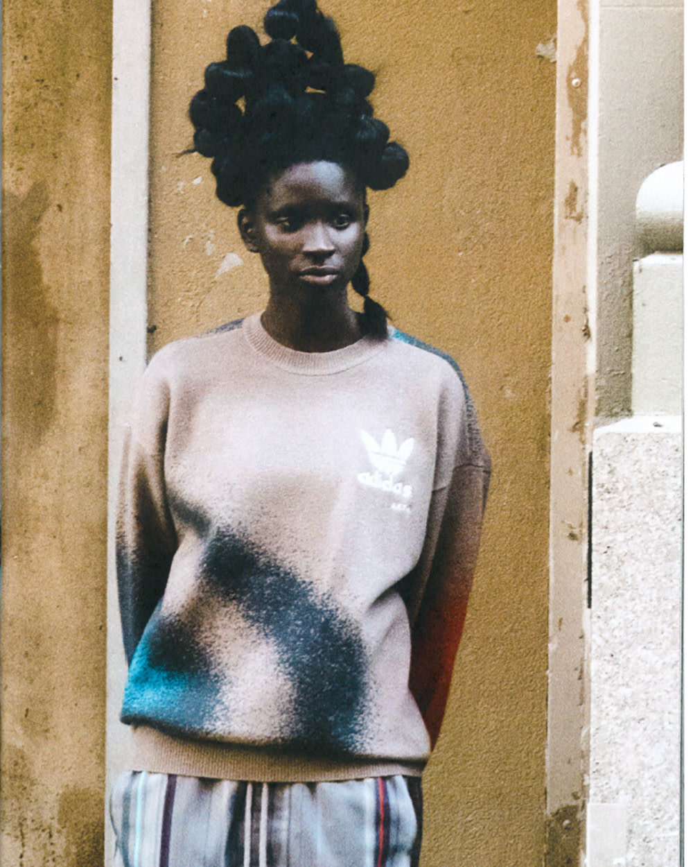 Woman in beige Adidas sweatshirt and striped pants poses against a textured, neutral wall. Hair is styled in sculptural buns.
