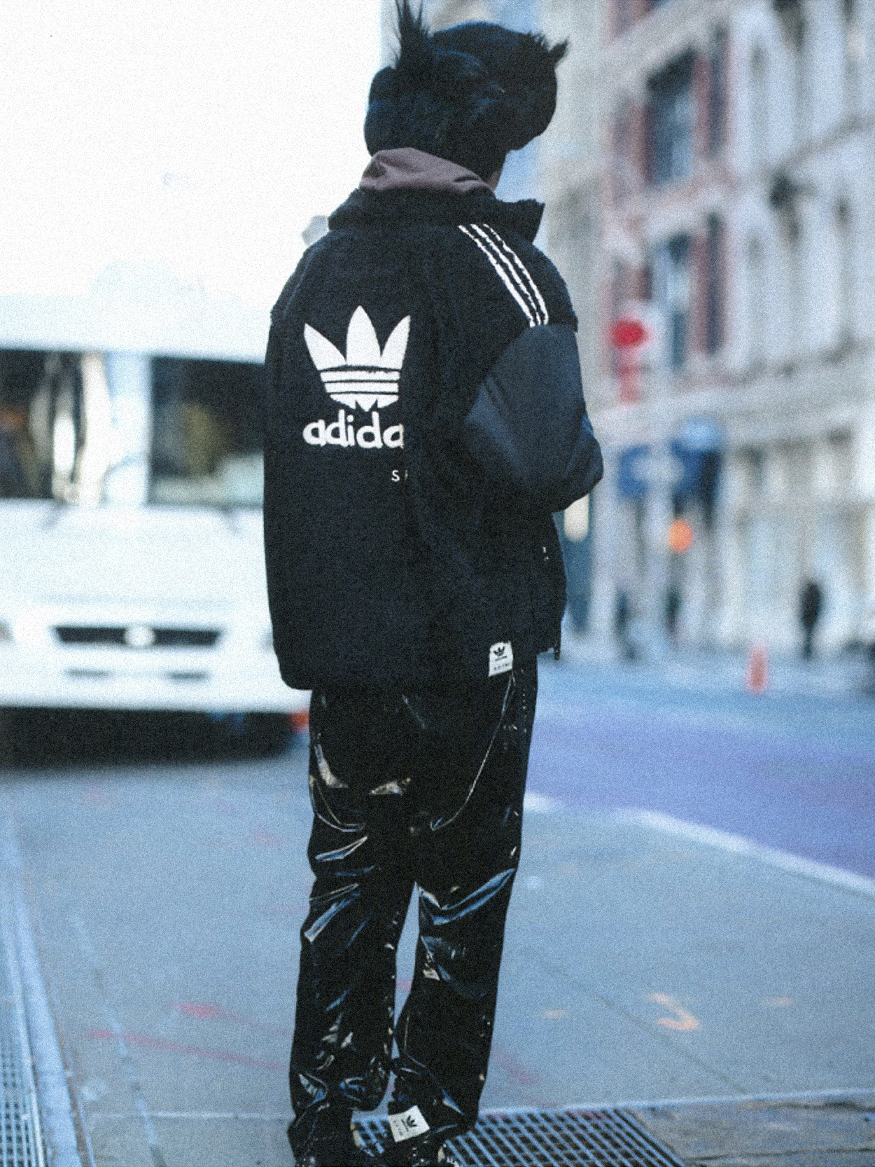 A person in an Adidas tracksuit, black furry hat, and shiny pants standing on a street with buildings and a vehicle in the background.