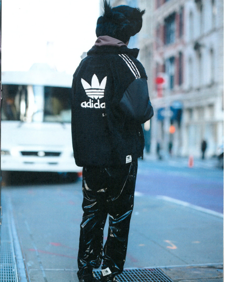 A person wearing a black Adidas jacket and shiny black pants stands on a city sidewalk with a blurry bus in the background.