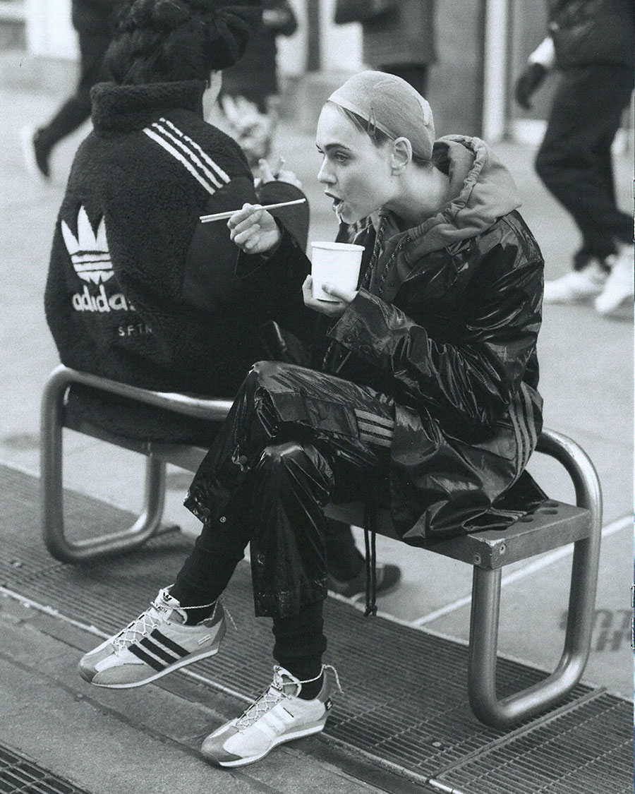 Woman in shiny black Adidas suit sits on bench, eating with chopsticks from white cup. Second person in jacket visible in background.