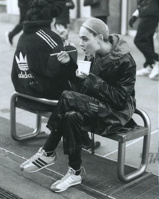 Woman in shiny black Adidas suit sits on bench, eating with chopsticks from white cup. Second person in jacket visible in background.