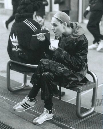 Woman in shiny black Adidas suit sits on bench, eating with chopsticks from white cup. Second person in jacket visible in background.