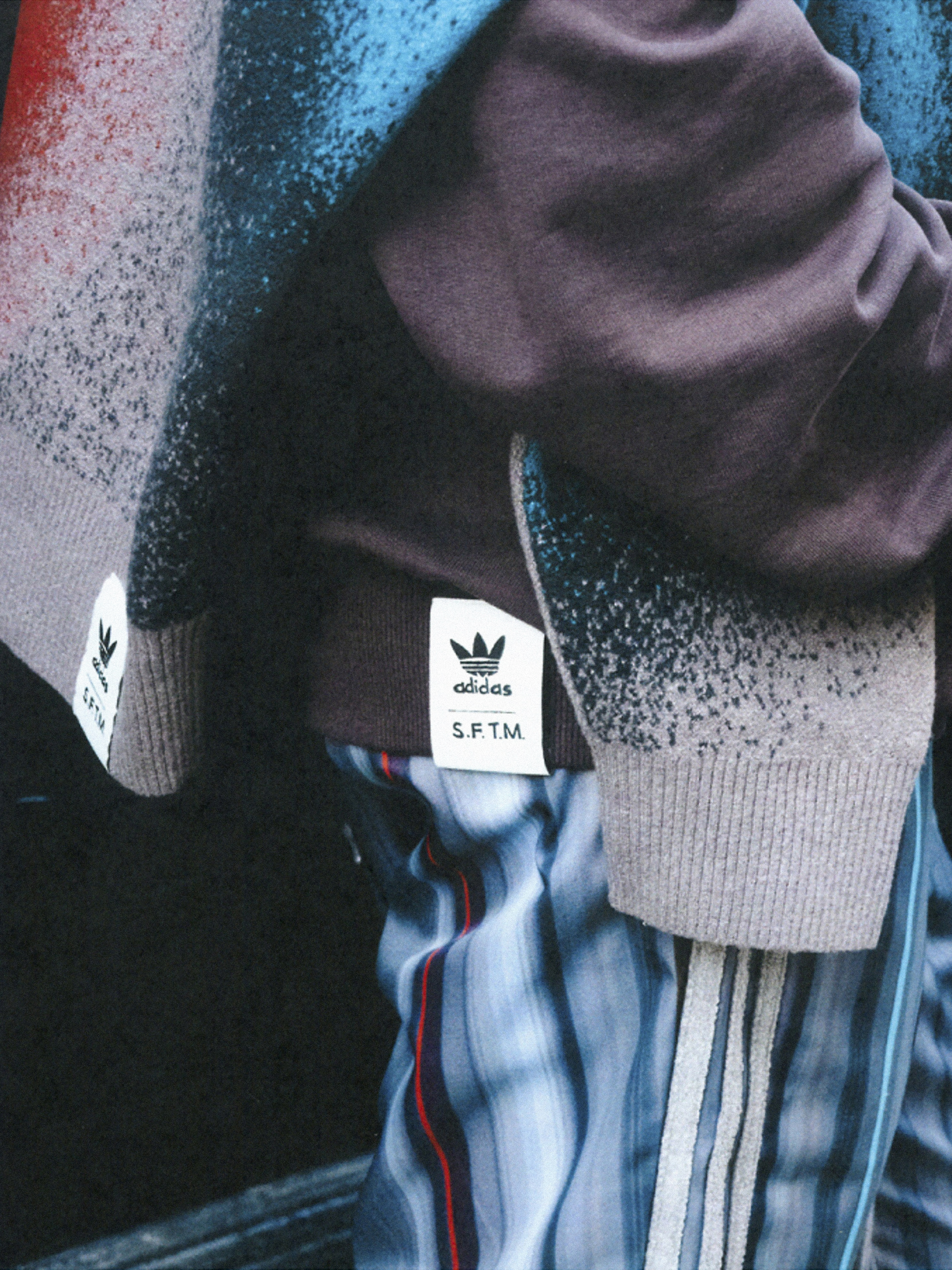 Close-up of person wearing Adidas apparel with gray and blue striped pants, a brown sweatshirt, and a black background. Apparel labels visible.