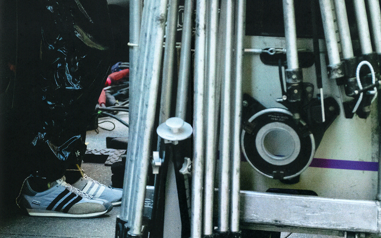 A low-angle shot captures a person wearing grey Adidas sneakers standing near metal scaffolding and film equipment on a film set.