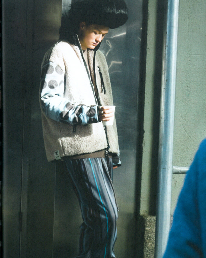 A person in a patterned jacket and striped pants holds a cup in an urban setting, with a metallic wall and handrail visible in the background.