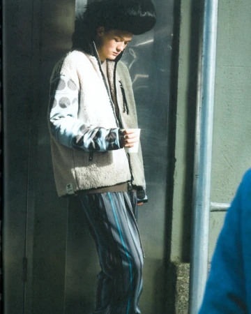 A person in a patterned jacket and striped pants holds a cup in an urban setting, with a metallic wall and handrail visible in the background.