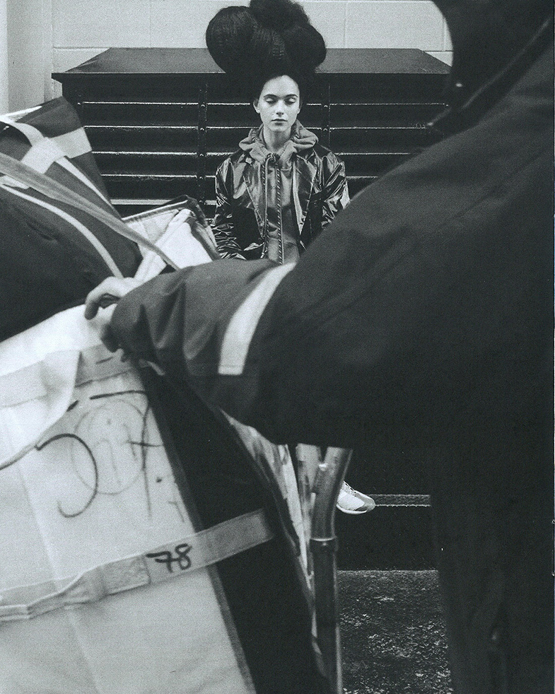 Monochrome fashion shot of a model in a glossy jacket and oversized updo standing behind mailbags and a postal worker in shadow.