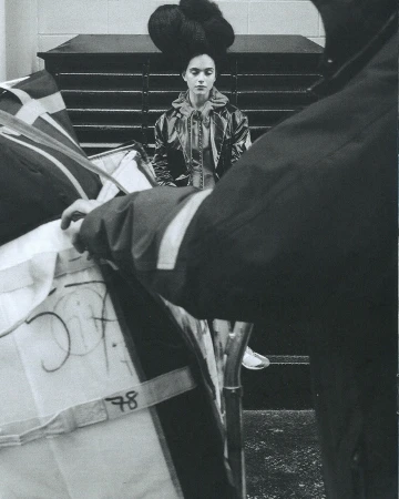 Monochrome fashion shot of a model in a glossy jacket and oversized updo standing behind mailbags and a postal worker in shadow.