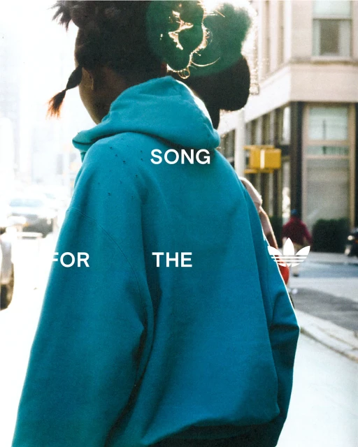 Person in teal hoodie walking on city sidewalk with Adidas logo. Back view. 'Song for the' text on back. Cars, buildings in background.