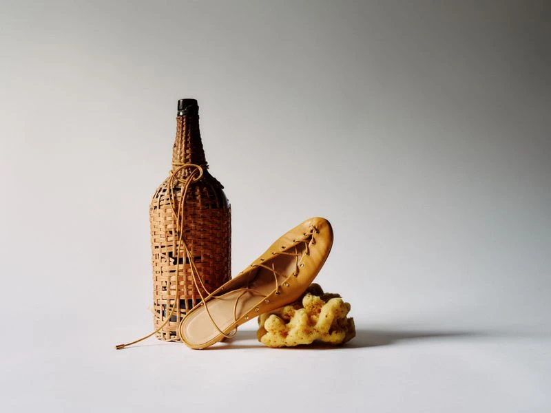 Fashion still life: tan-colored lace-up shoe on a sea sponge next to a decorative wicker bottle, neutral gradient background.