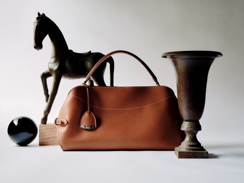 Fashion still life: a tan leather handbag is centered in the frame, flanked by a dark horse sculpture and decorative vase, creating an elegant composition.