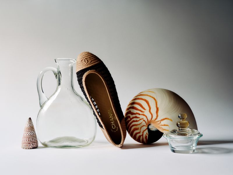 Still life featuring a Chanel shoe, nautilus shell, glass objects, and a small woven cone against a gradient grey background.