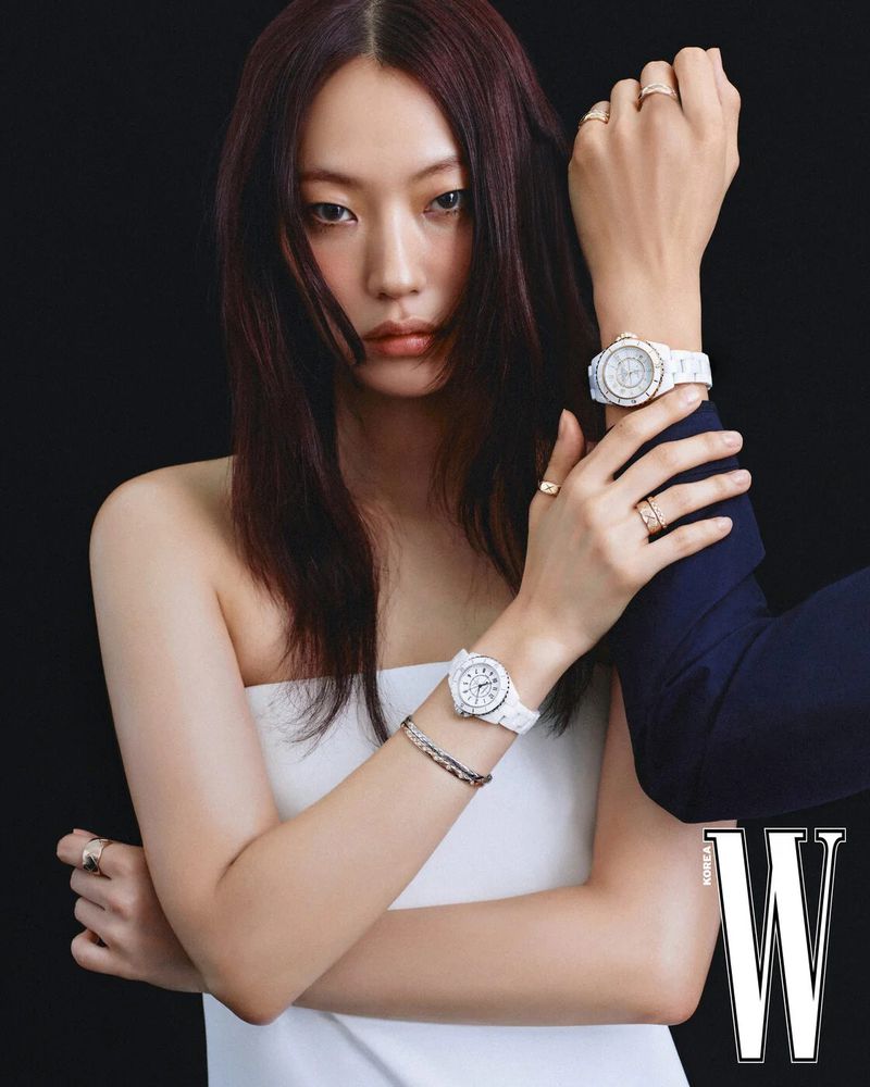 Close-up of Asian woman with dark hair wearing a strapless white dress and white ceramic watch and gold rings, dark background.