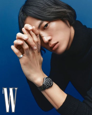 Close-up of an adult male with dark hair and a black turtleneck posing against a blue background, wearing a wristwatch and ring.