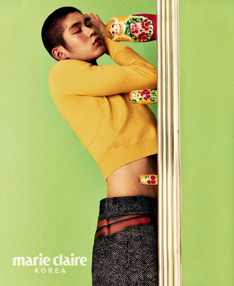 An Asian male model wearing a yellow top and dark pants poses with Russian dolls against a green backdrop in a fashion editorial shot.
