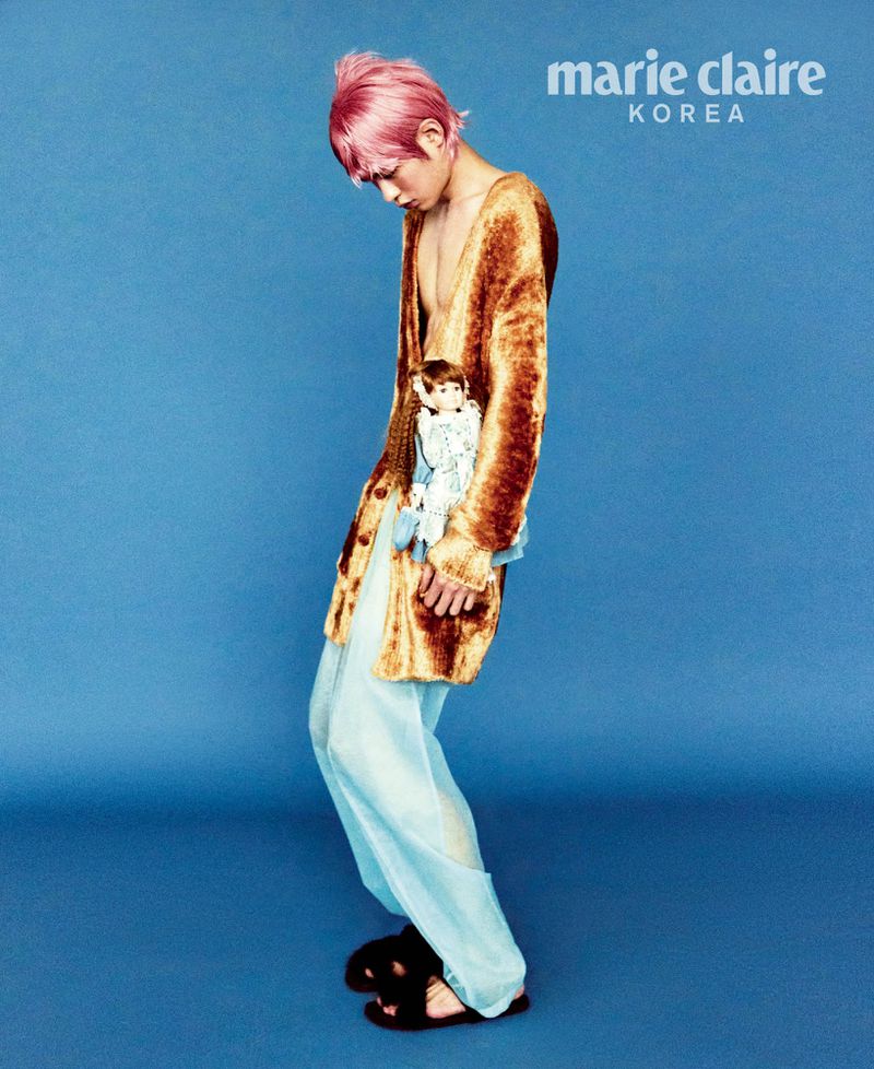 A person with pink hair, in a gold robe and blue pants, holds a doll in a studio portrait against a blue backdrop for Marie Claire Korea.