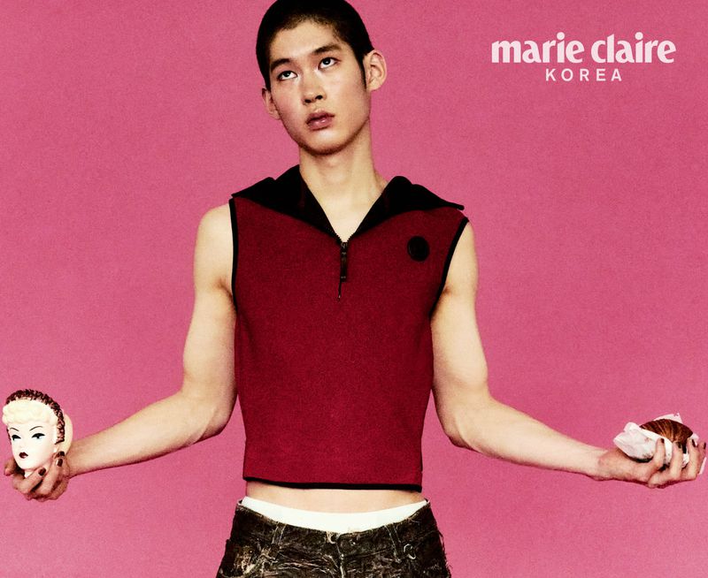 A young Asian male model poses in a sleeveless top and patterned pants against a pink background, holding a baked good in each hand.