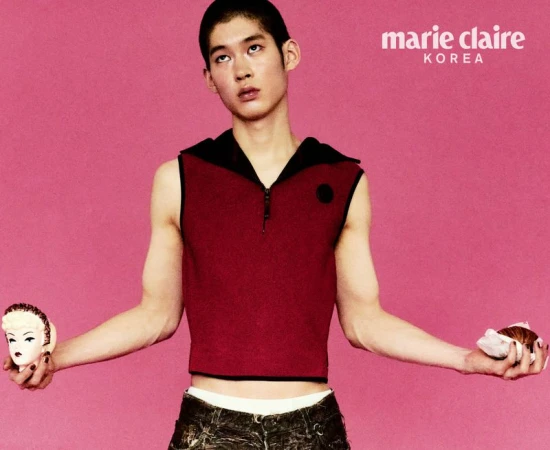 A young Asian male model poses in a sleeveless top and patterned pants against a pink background, holding a baked good in each hand.