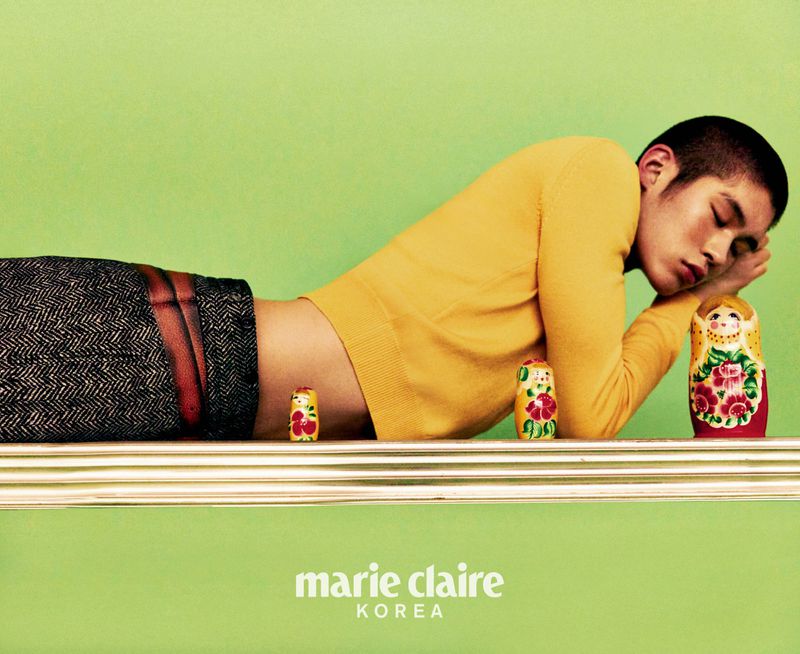 Stylish adult male with short hair, lying on shelf with eyes closed next to set of three nesting dolls in a studio with green backdrop.