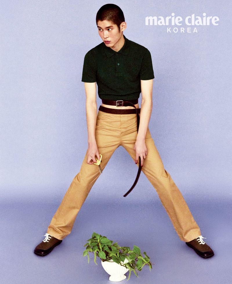Editorial fashion shot of a young adult male model with buzzcut, wearing dark green shirt and khaki pants, standing in a light purple studio.