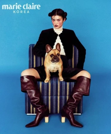 A woman in a black jacket and brown leather boots sits on a striped chair with a French bulldog in a blue studio setting.