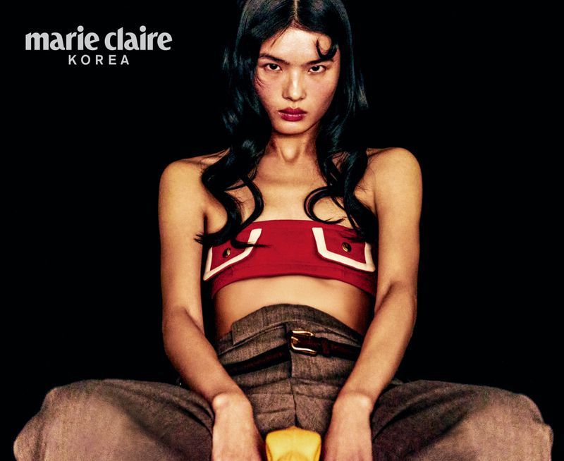 Fashion editorial shot featuring an Asian woman in a red top and gray pants, seated against a dark background, holding a yellow object.