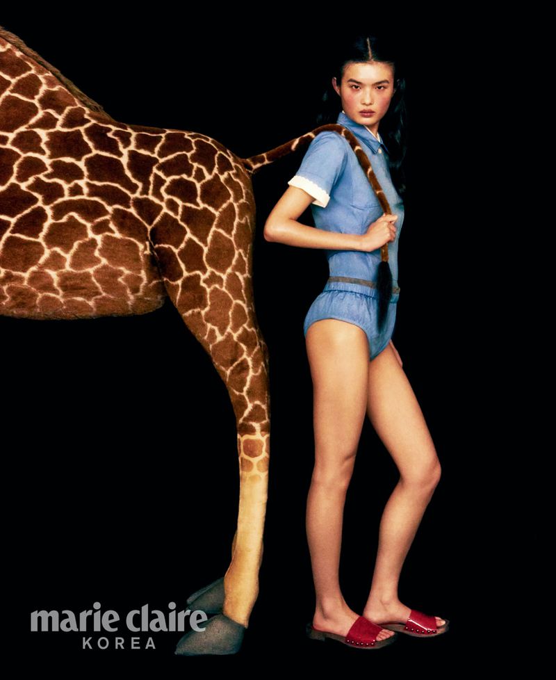 Fashion shot of a woman in a light blue outfit posing next to a giraffe's rear on a black background for Marie Claire Korea magazine.