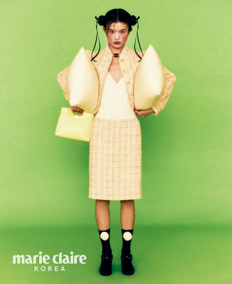 Editorial fashion shot of an Asian model in a yellow skirt suit holding two pillows and a bag against a vibrant green background.