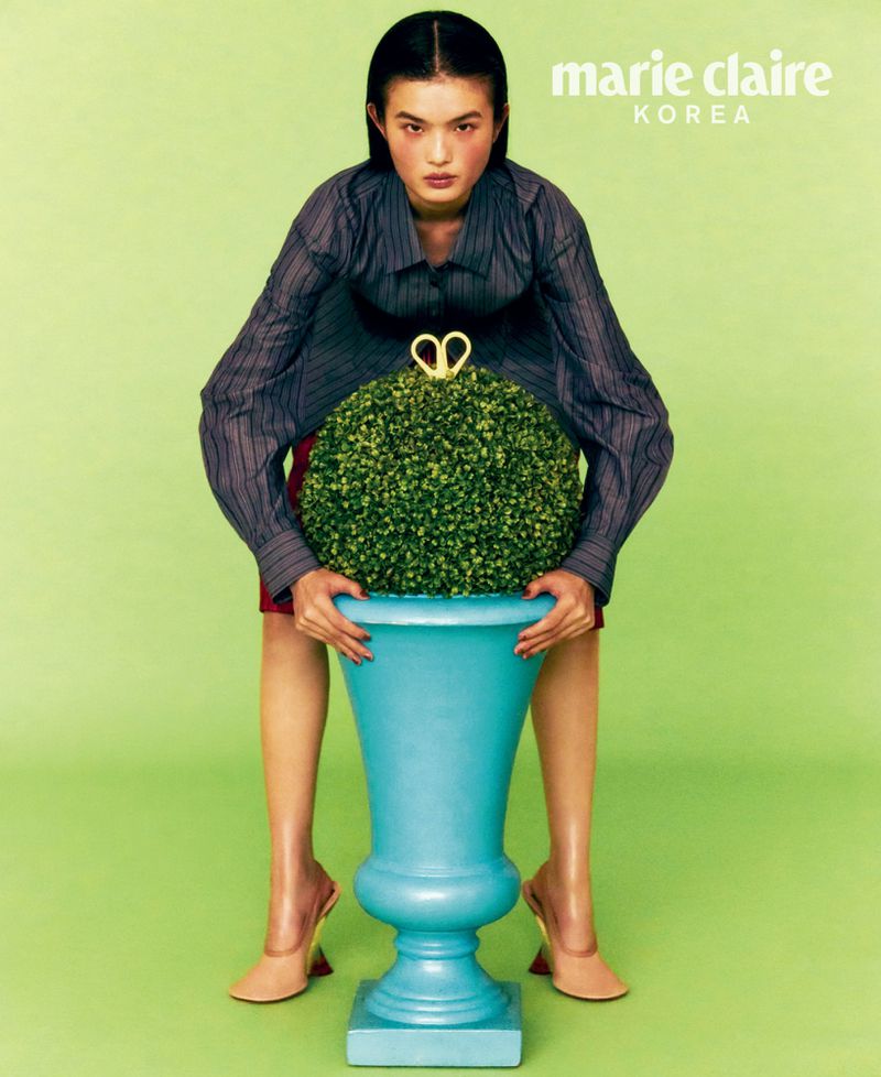 Editorial fashion shot of a woman holding a blue planter with a topiary against a lime-green backdrop, wearing a striped shirt and heels.