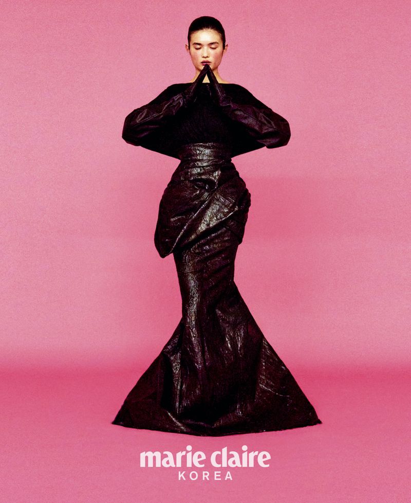A fashion shot of a woman in a black textured gown against a pink background, hands clasped in front of her chest, editorial.