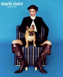 Woman with space buns seated on an armchair with a French bulldog, wearing a black jacket and tall boots against a blue background.