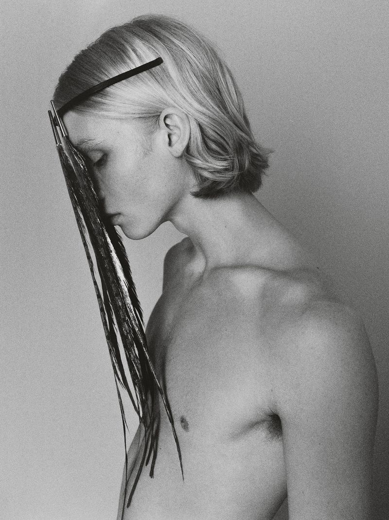 Artistic portrait of a bare-chested young adult male with blond hair, a black headband, and face partially obscured by black feathers.