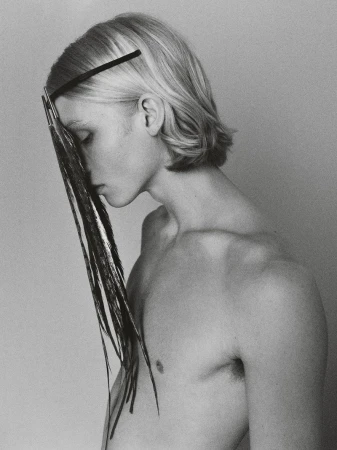 Artistic portrait of a bare-chested young adult male with blond hair, a black headband, and face partially obscured by black feathers.