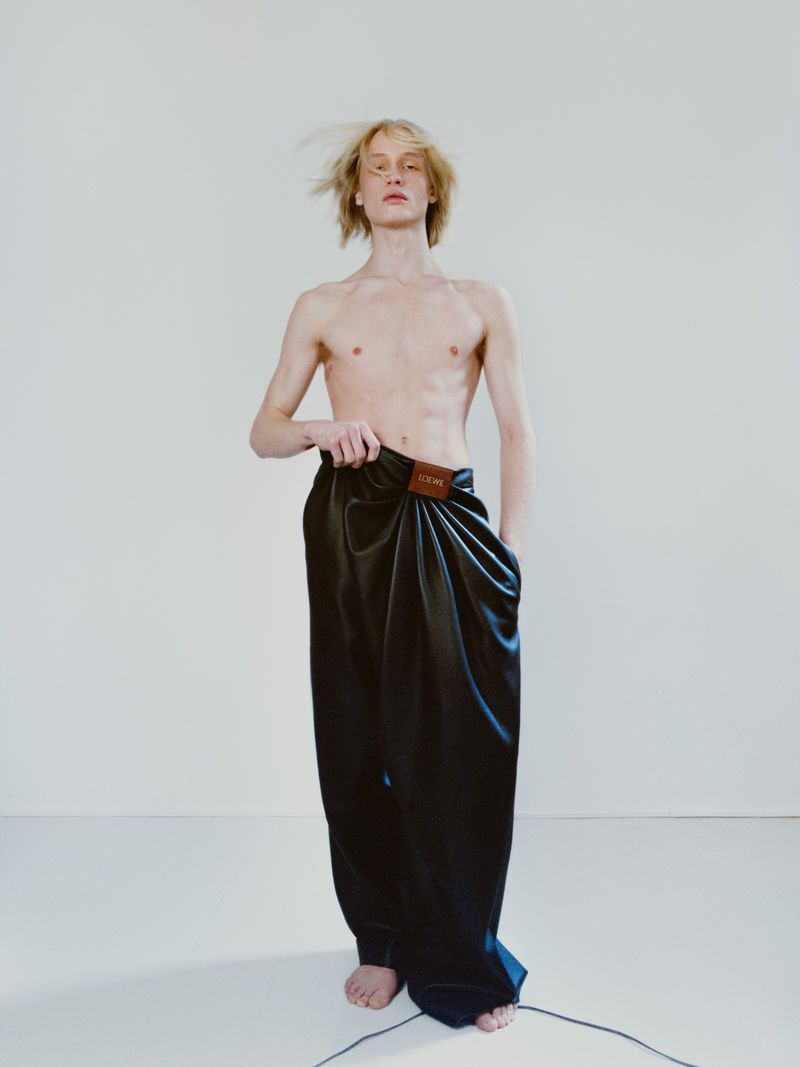 Adult male fashion model poses shirtless in draped black pants with a logo patch, against a white background in a studio setting.