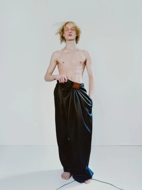 Adult male fashion model poses shirtless in draped black pants with a logo patch, against a white background in a studio setting.