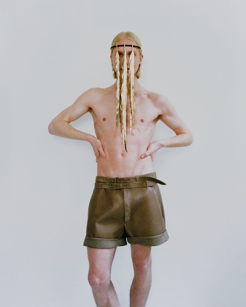 Fashion shot of a shirtless blonde male model in shorts with a decorative headpiece, posing against a stark white backdrop.