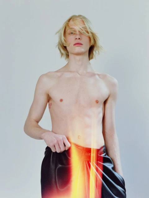 Close-up studio shot of a shirtless male model with blond hair wearing black pants with a blurred flame pattern. Fashion editorial.