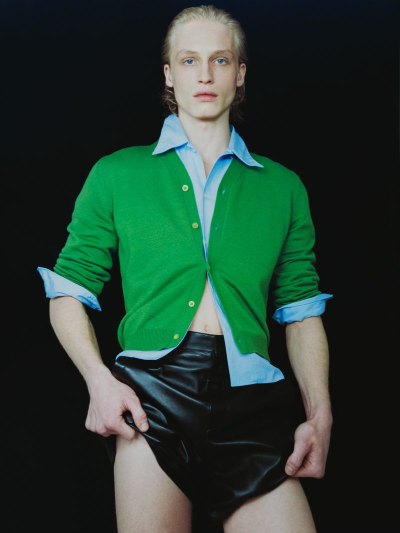 Young adult man in a green cardigan and black leather shorts poses against a dark background in a fashion editorial style shot.